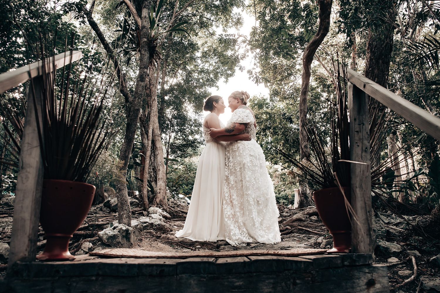 A bride in a white wedding dress stands on a wooden platform in a lush, forested environment.