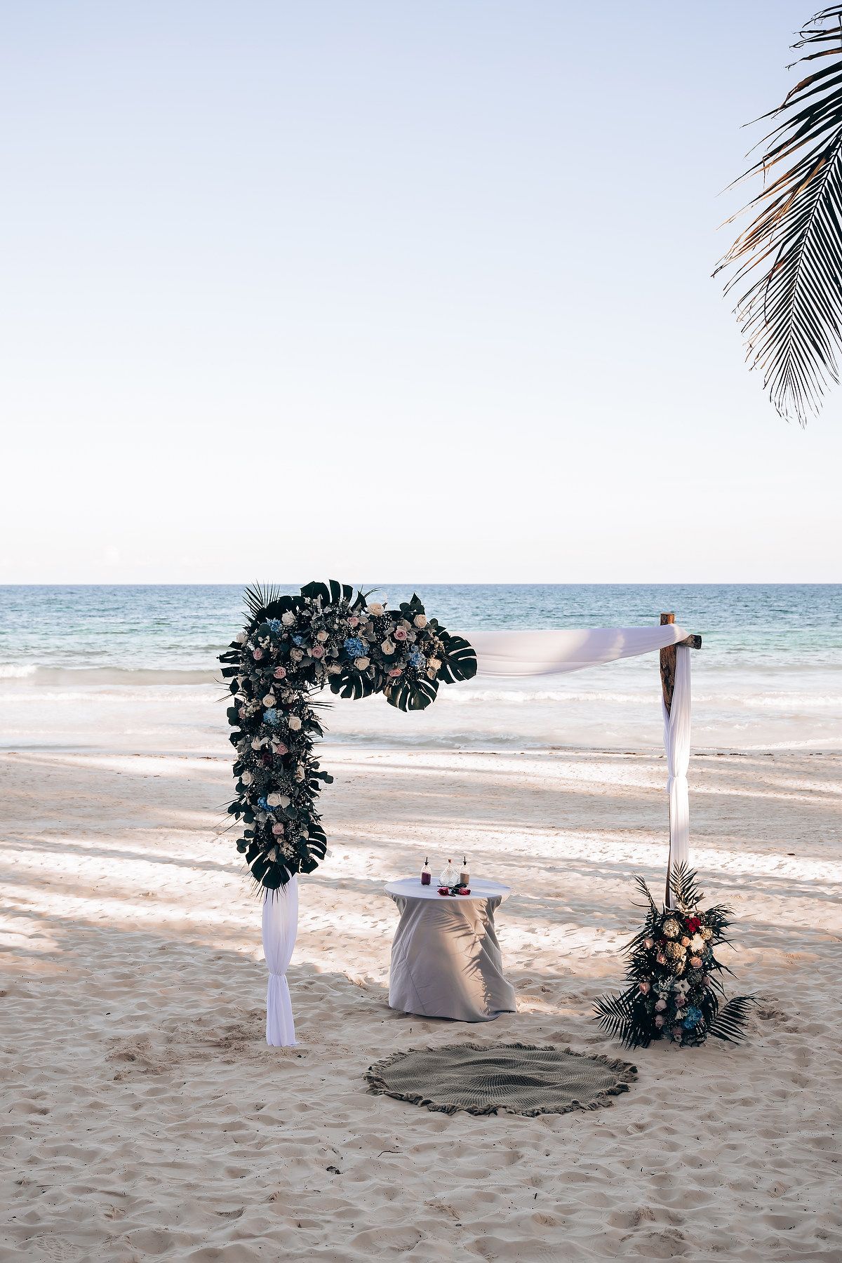The image shows a tropical beach scene with a palm tree and a wedding arch decorated with flowers on the sand.