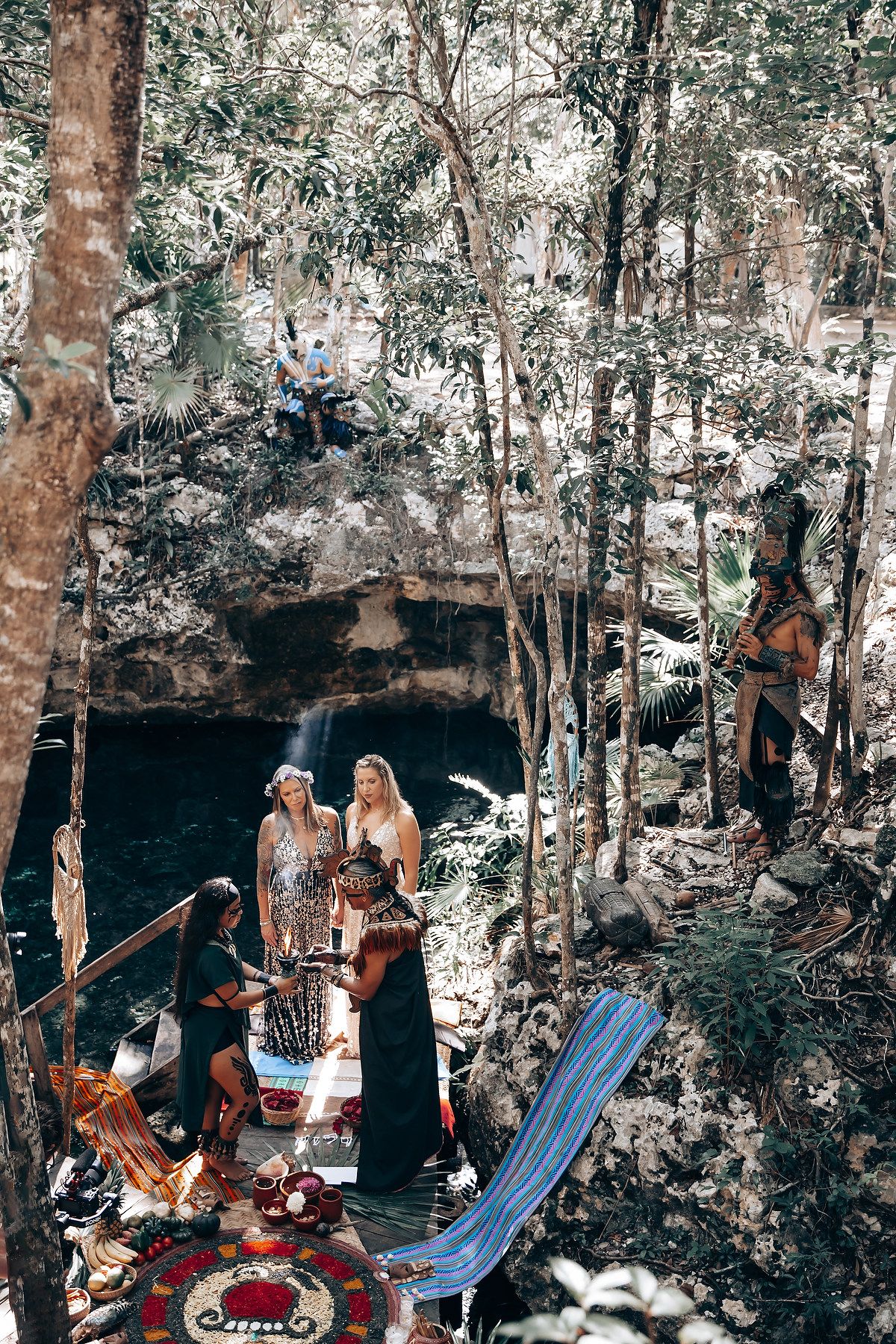 A group of people standing near a natural pool or spring in a wooded area, with sunlight and vegetation visible.
