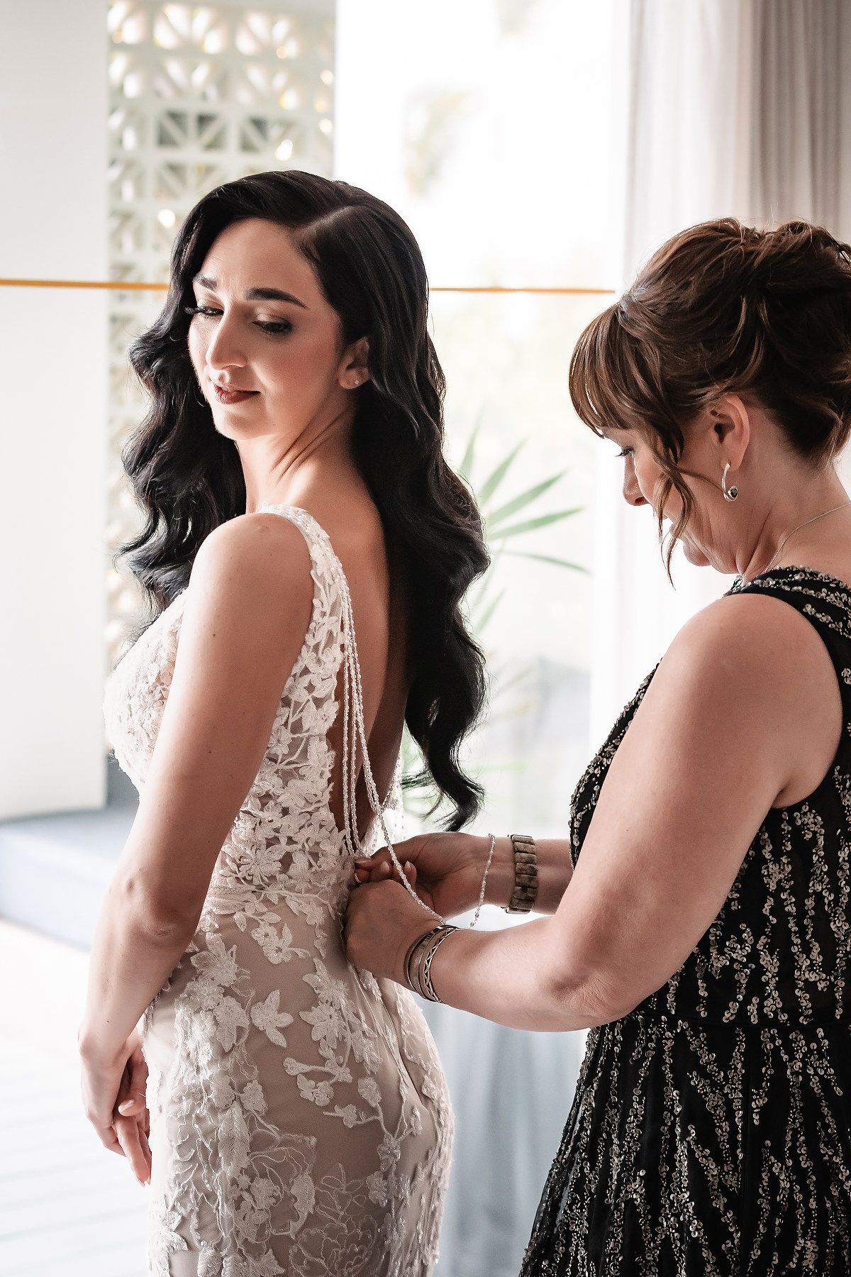 Mother helping bride fasten her lace wedding dress from behind in a bright elegant room.