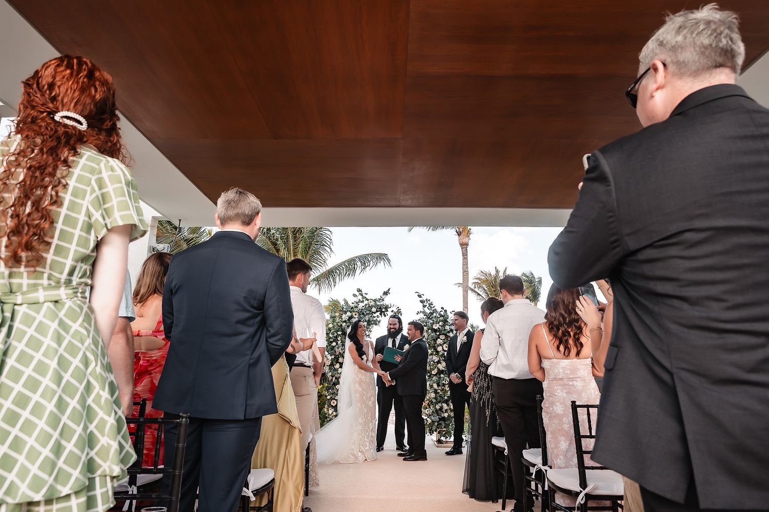 Outdoor wedding ceremony with guests standing and watching couple exchange vows under a covered pavilion near tropical palm trees.
