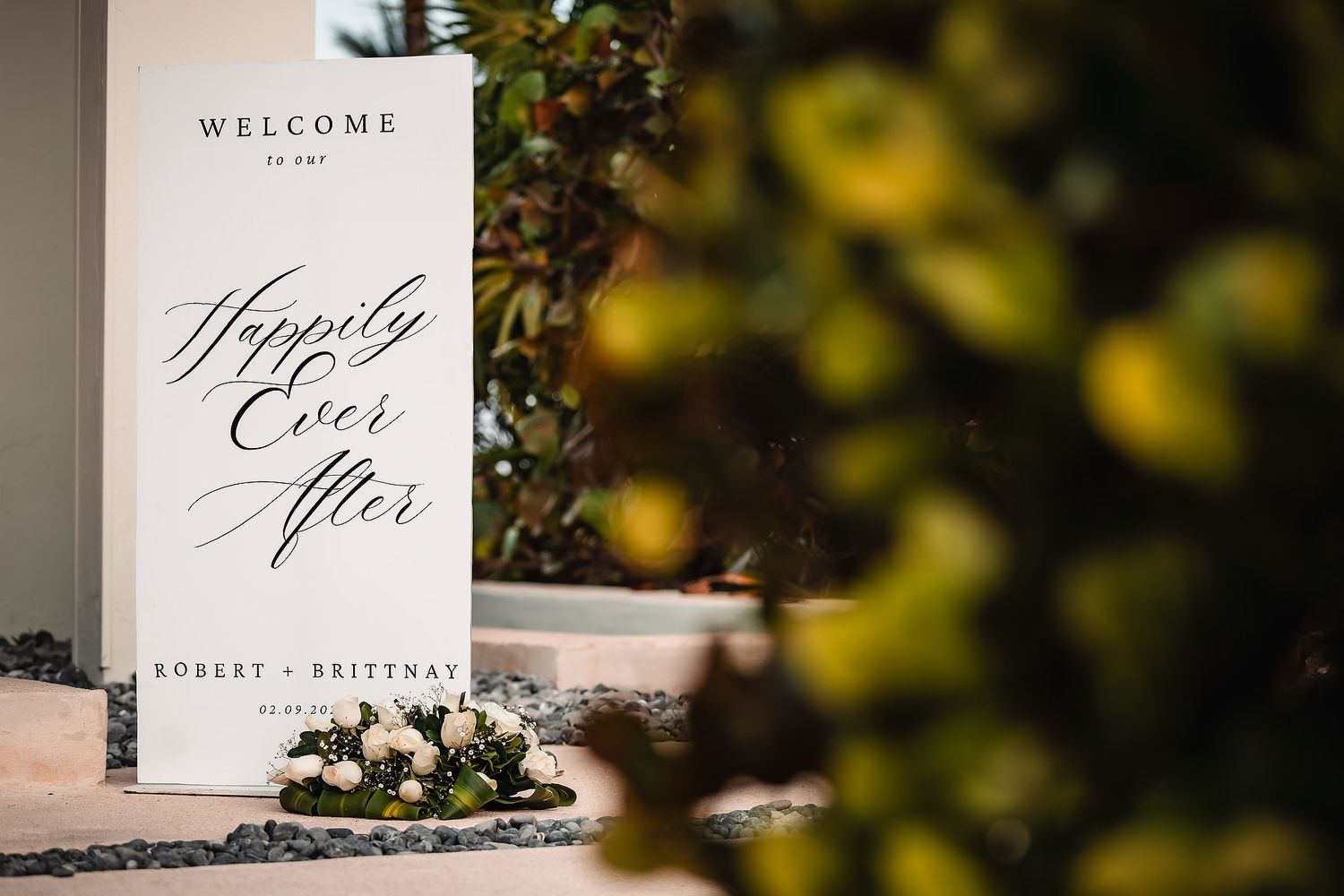 Elegant wedding welcome sign reading Happily Ever After for Robert and Brittnay, framed by lush greenery and white flowers.