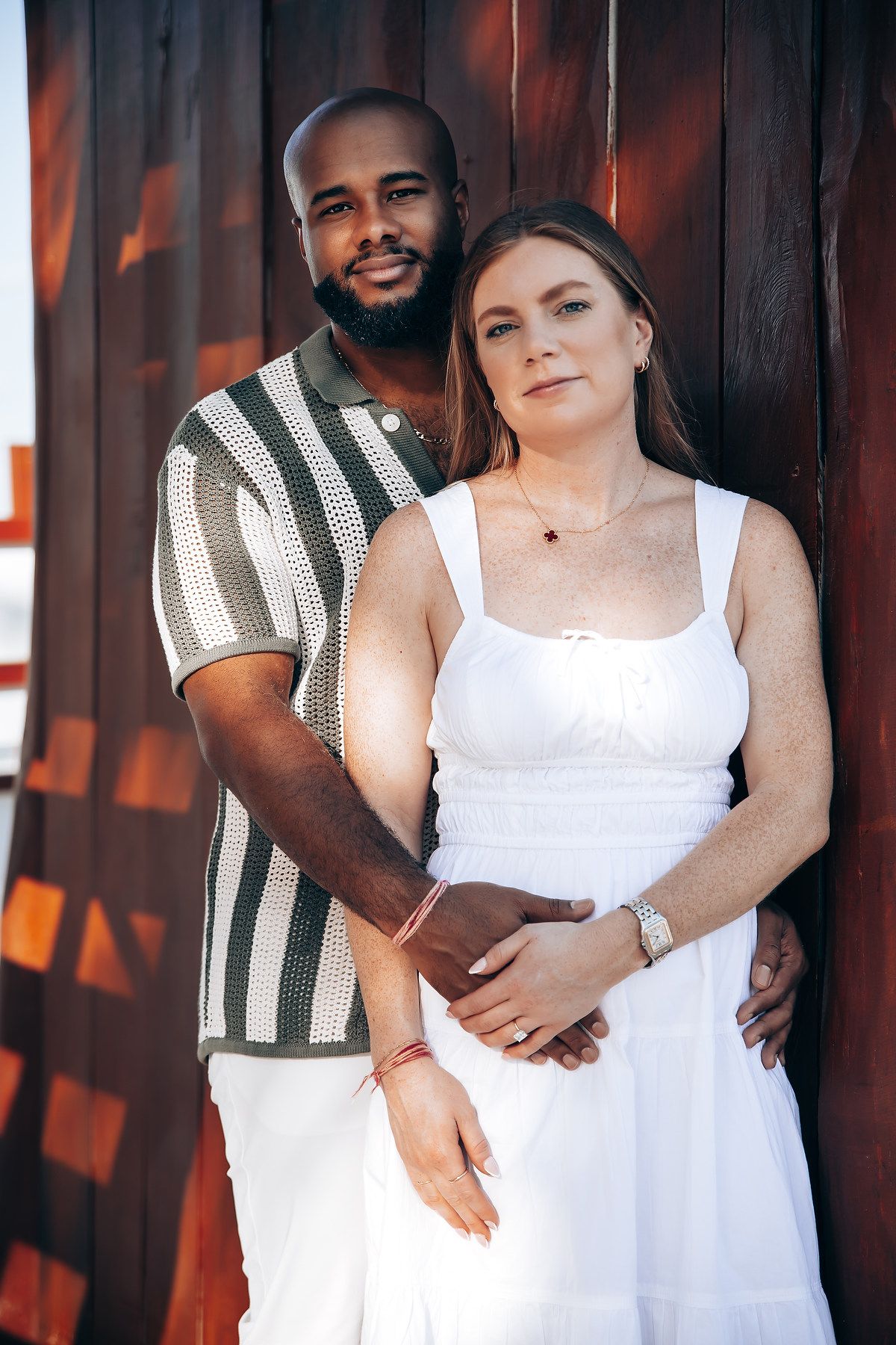 Interracial couple posing closely against a warm wooden wall, woman in white dress, man in striped shirt.