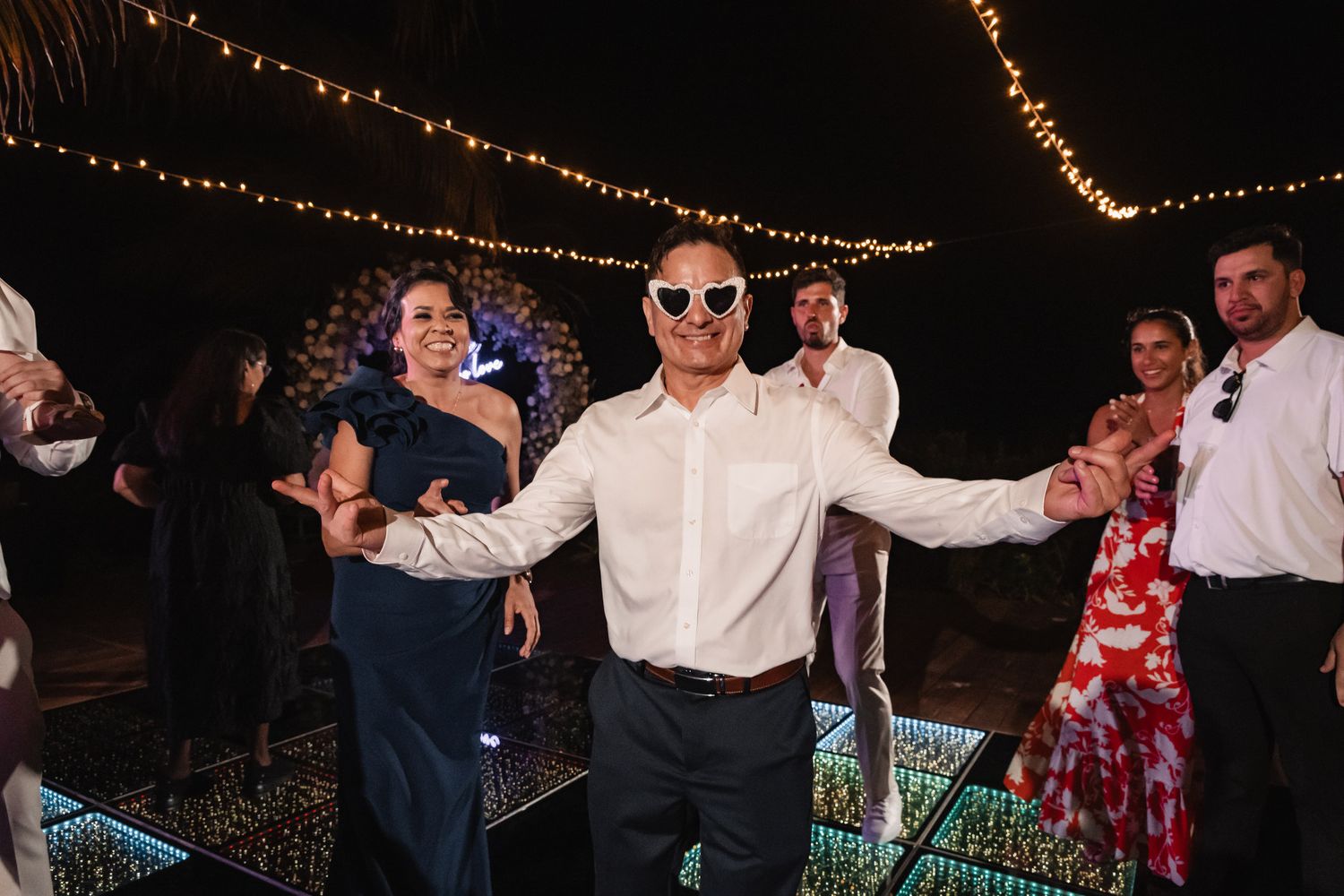 Wedding guest dancing in novelty sunglasses on illuminated dance floor during lively outdoor reception.