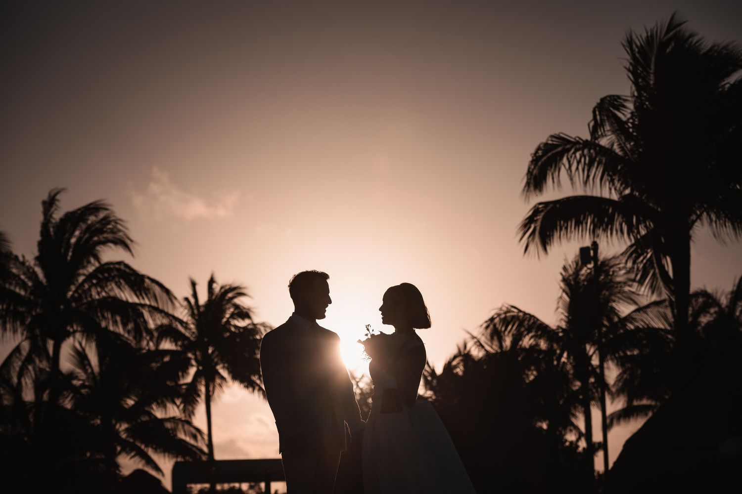 Silhouette of a bride and groom at sunset with palm trees at a tropical wedding venue.