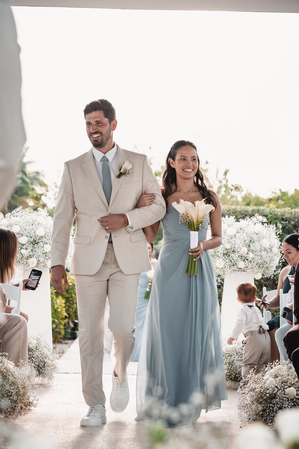 Smiling groomsman in beige suit escorting bridesmaid in blue gown down a flower-lined aisle.