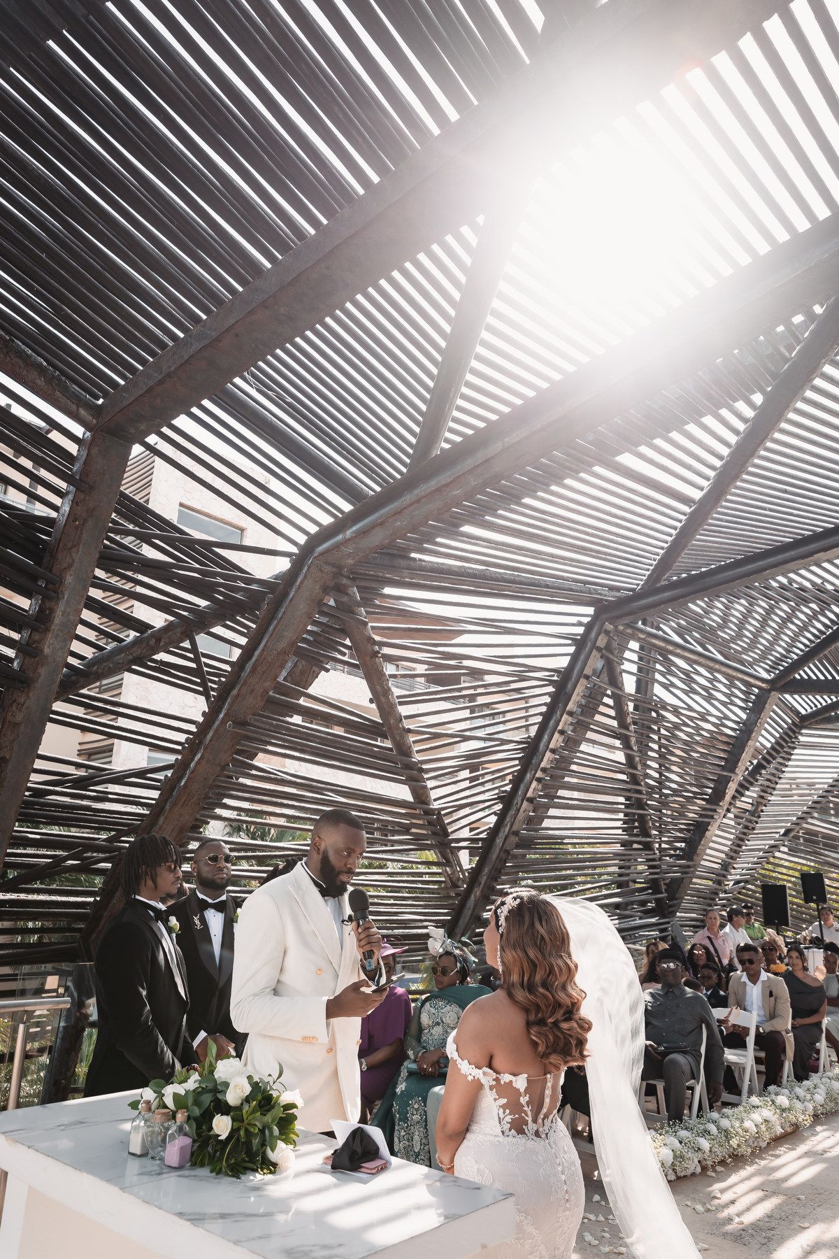 Groom in white suit exchanges vows with bride under a stunning wooden lattice ceremony structure.