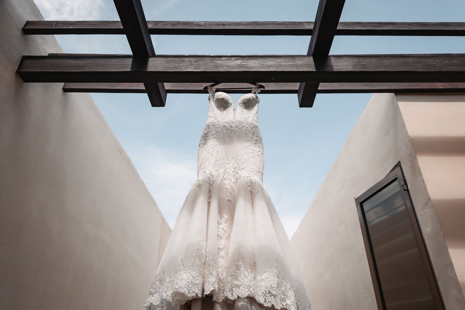 Stunning white lace bridal gown displayed hanging under a glass pergola with blue sky backdrop.