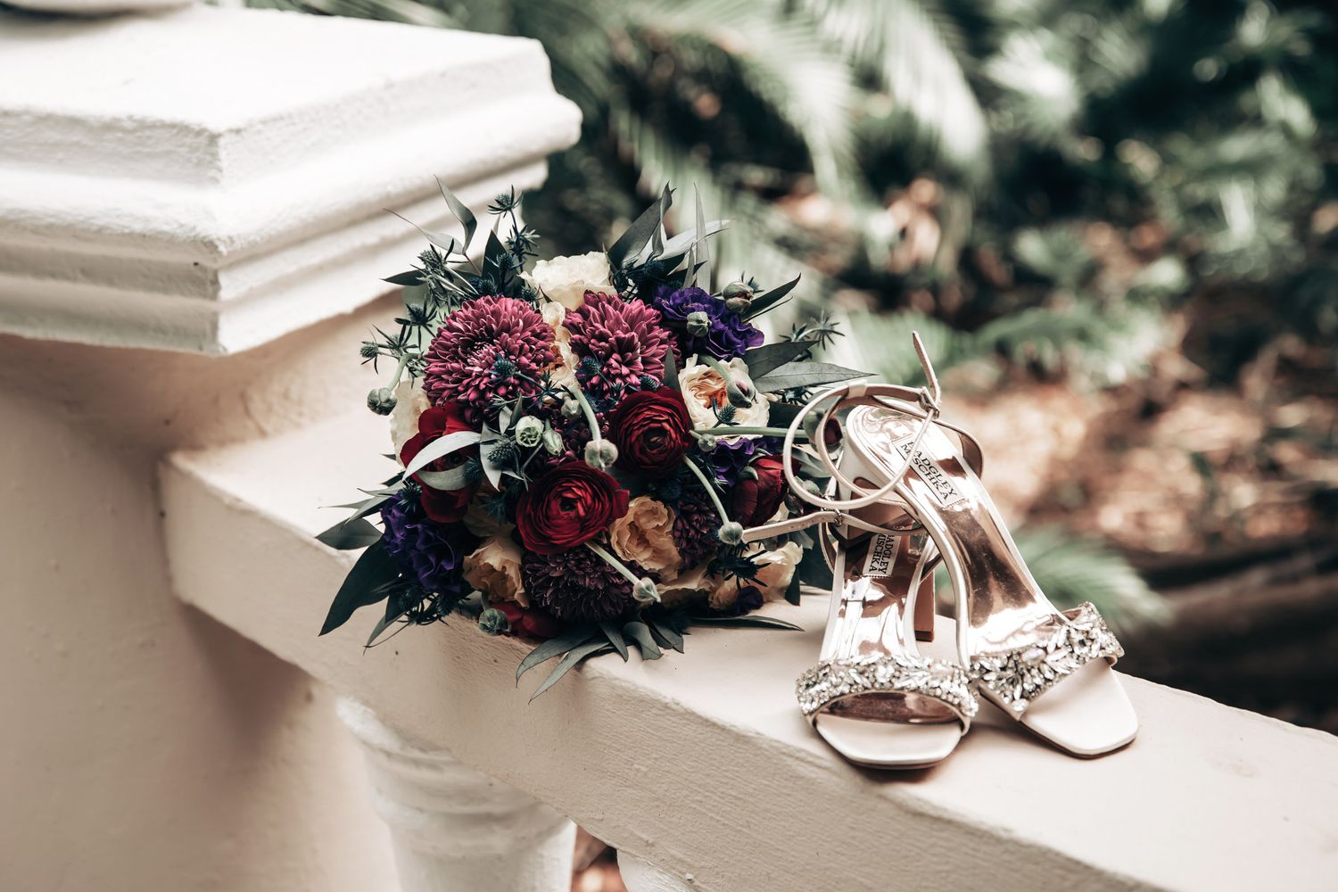 Bridal bouquet with burgundy and purple flowers beside embellished wedding heels on white railing.