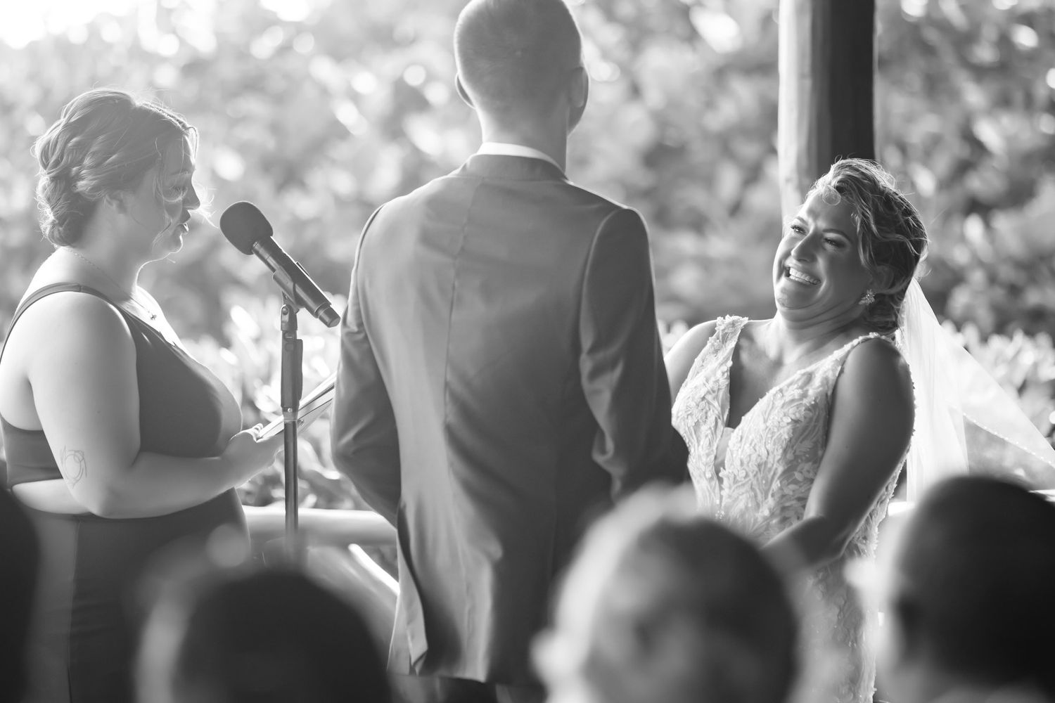 Black and white photo of bride smiling at groom during outdoor wedding ceremony with officiant at microphone.