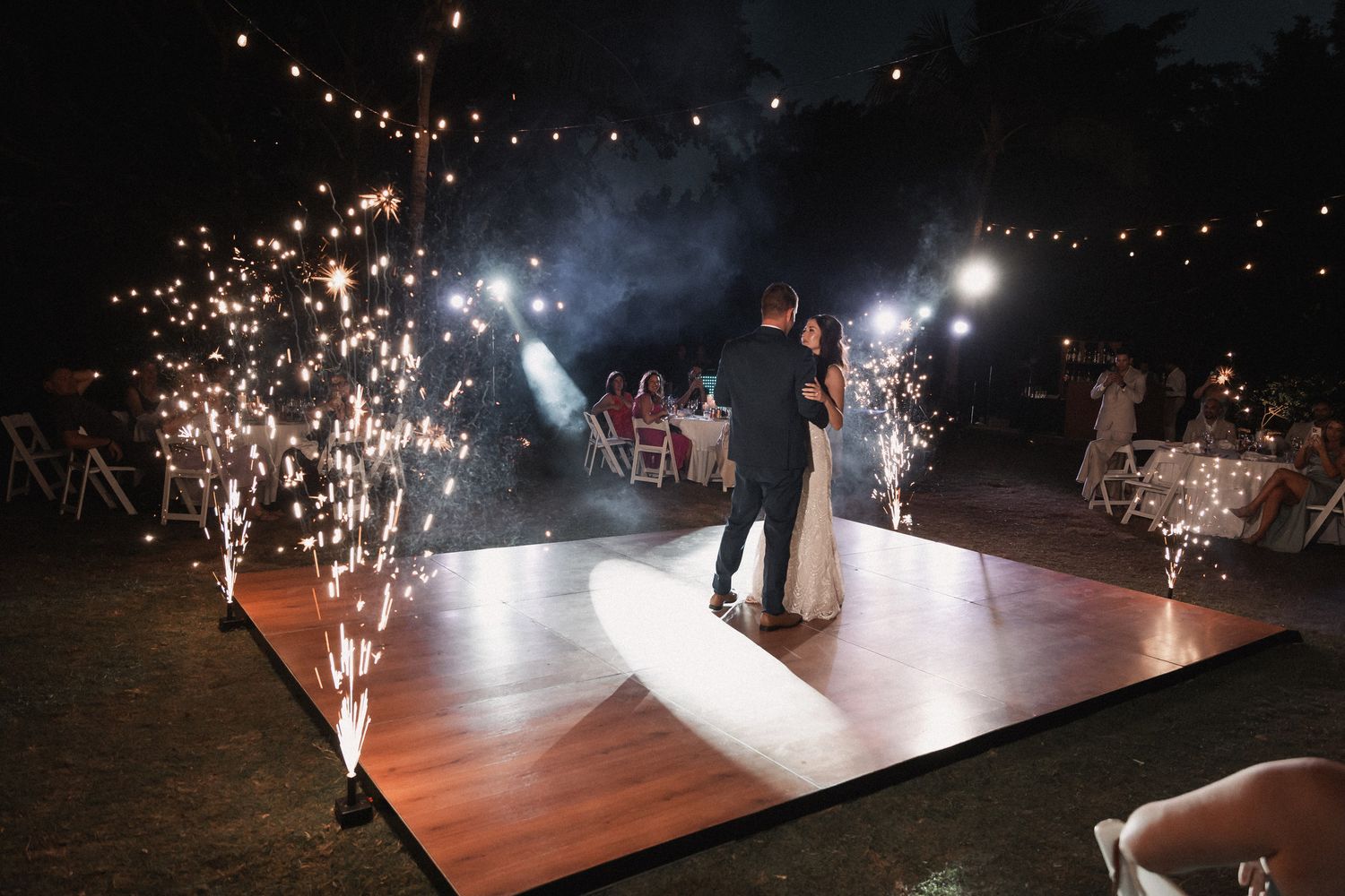 Bride and groom share first dance on illuminated floor surrounded by sparkling cold fireworks at night.
