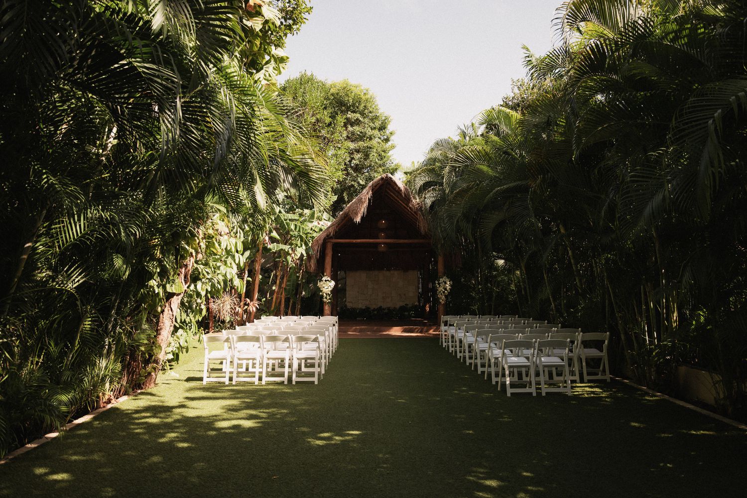 Outdoor tropical wedding ceremony setup with white chairs on green lawn leading to thatched roof altar.