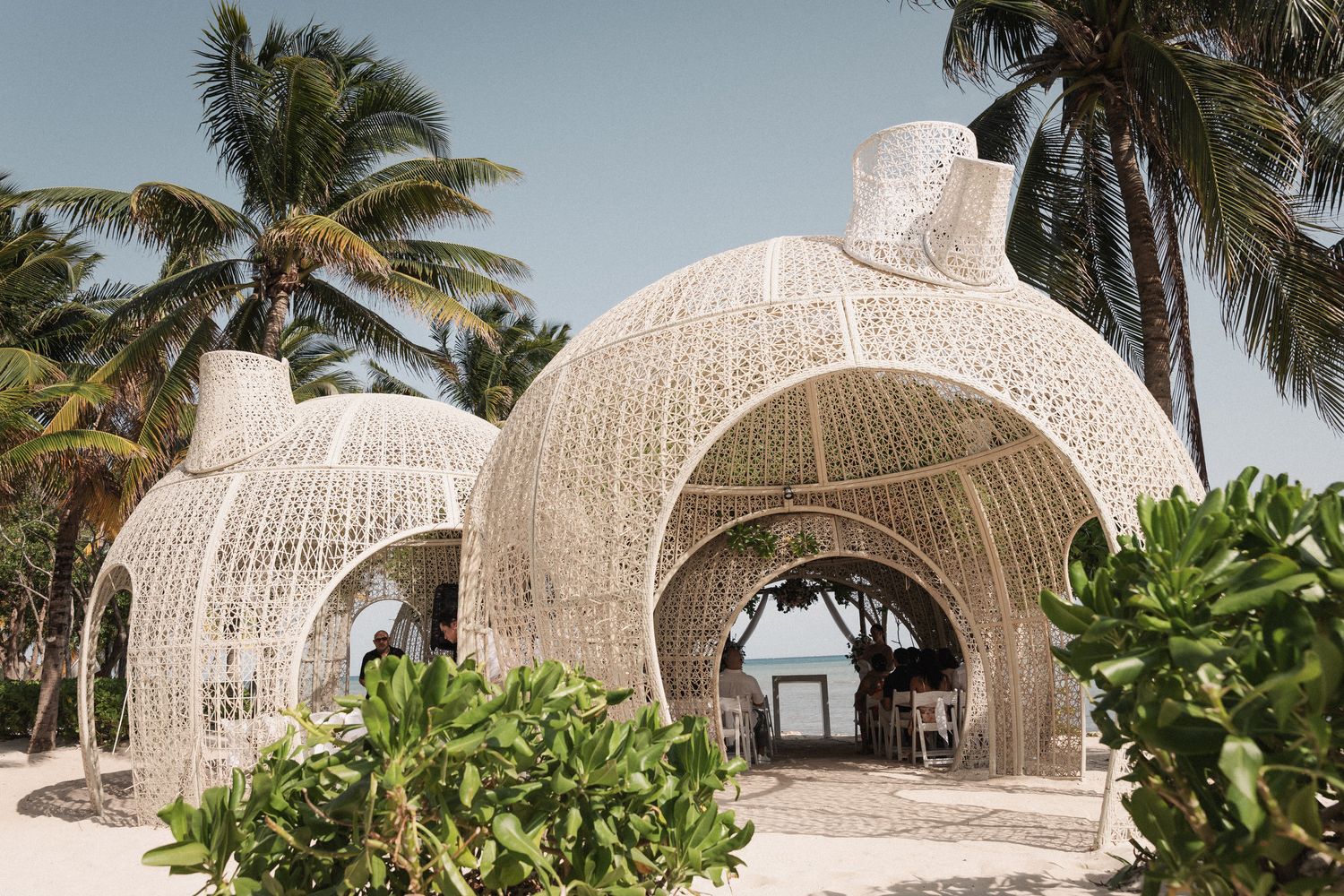 Woven bamboo dome structures on a tropical beach surrounded by palm trees and lush greenery.