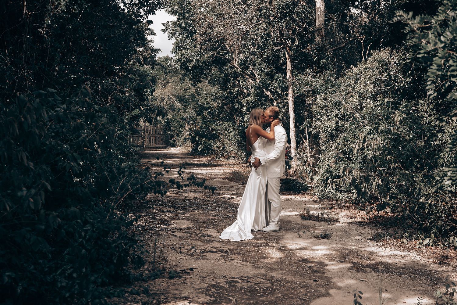 A couple embraces on a forest path surrounded by lush greenery and wearing wedding attire.