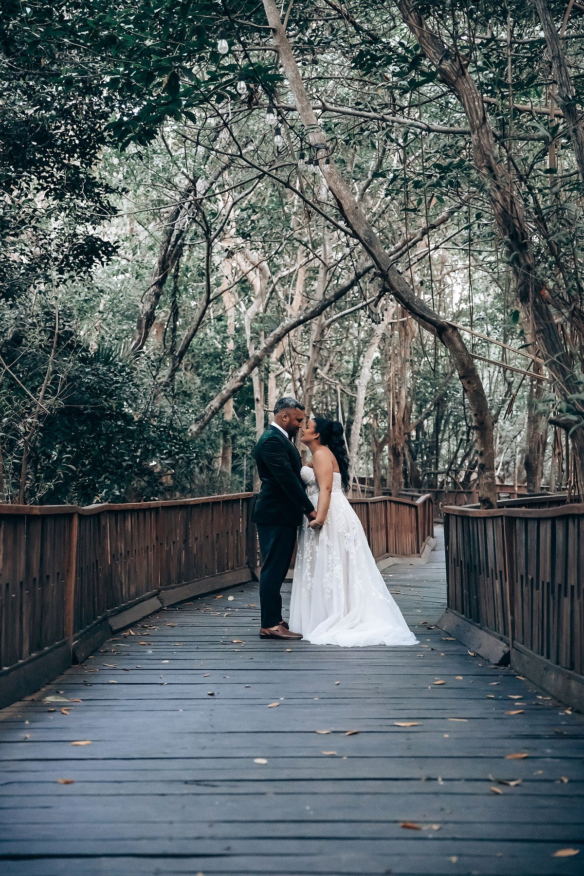 A couple in wedding attire share an intimate moment on a wooden boardwalk surrounded by lush forest trees.