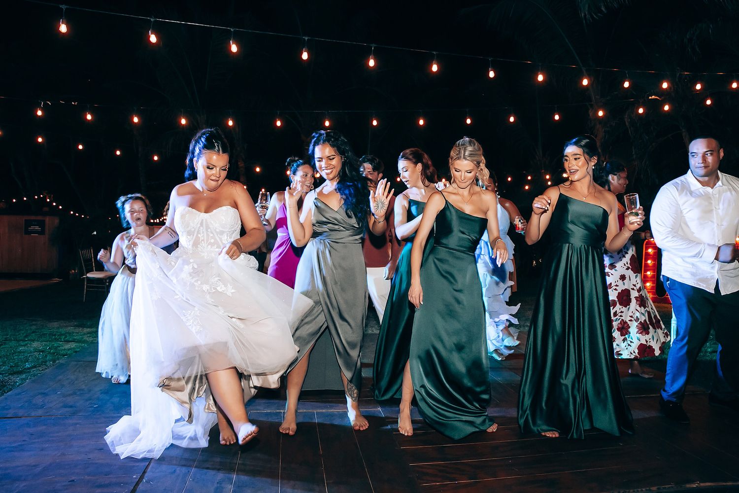 Wedding guests celebrate on outdoor dance floor under string lights at evening reception.