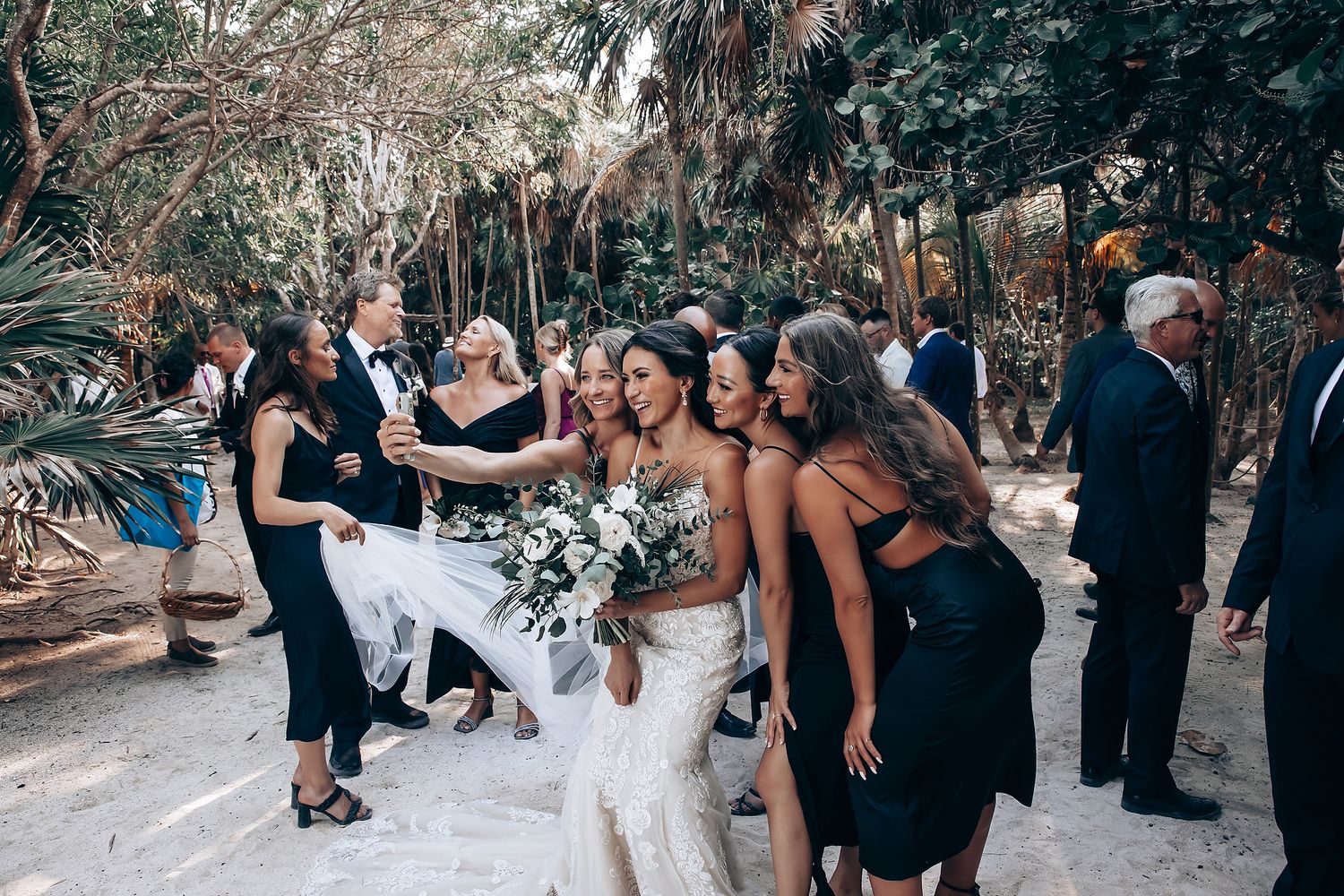 Wedding party gathered outdoors for group photos among tropical palm trees and greenery.