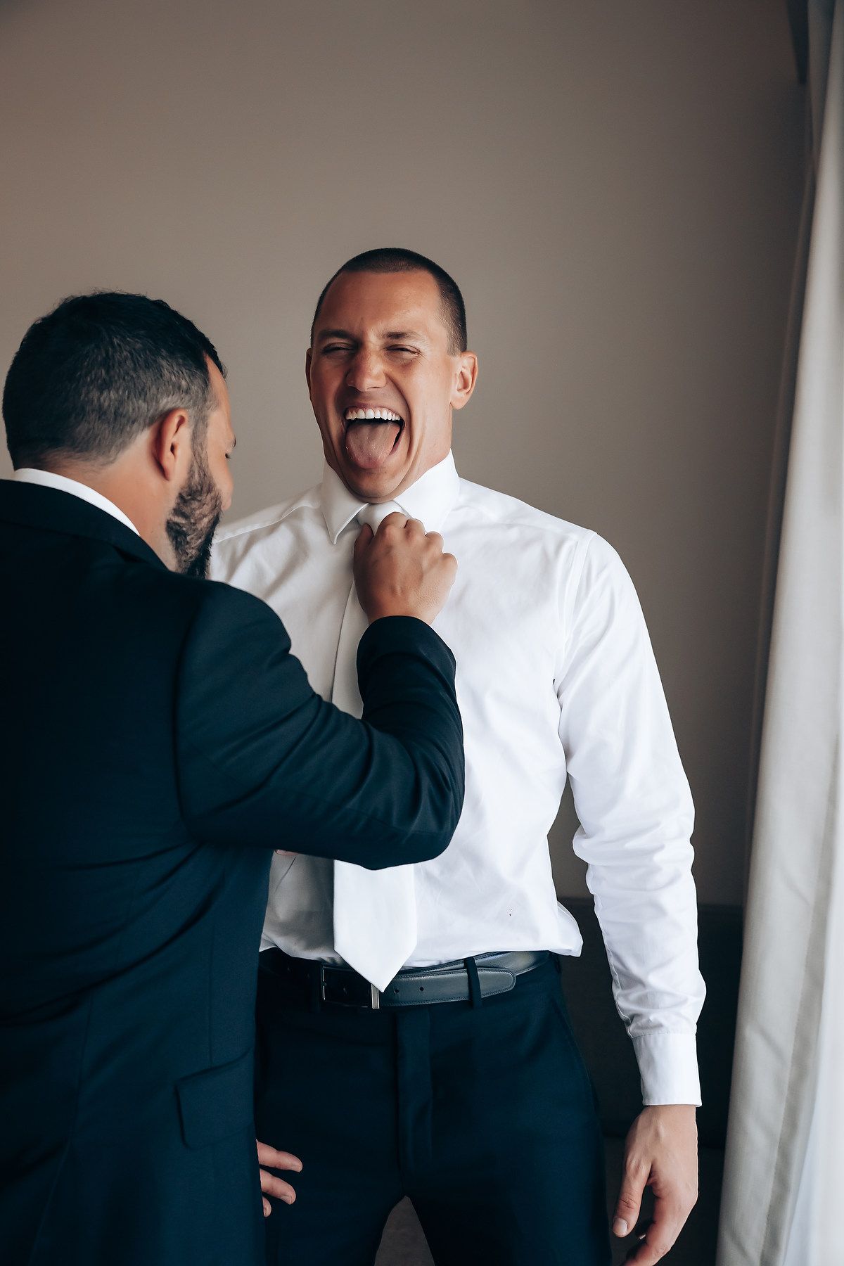 Two people share a candid laughing moment during wedding preparations in formal attire.