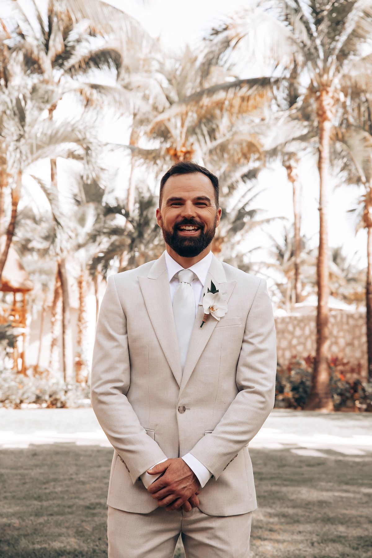 Groom in a beige suit poses for portraits in a tropical garden setting with palm trees.