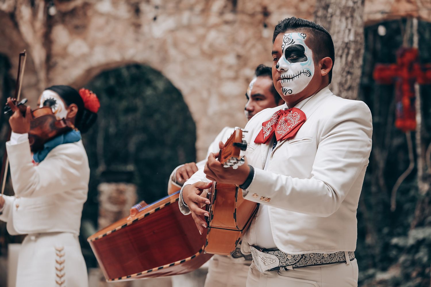 Mariachi musicians in traditional white suits and Day of the Dead face paint perform with instruments outdoors.