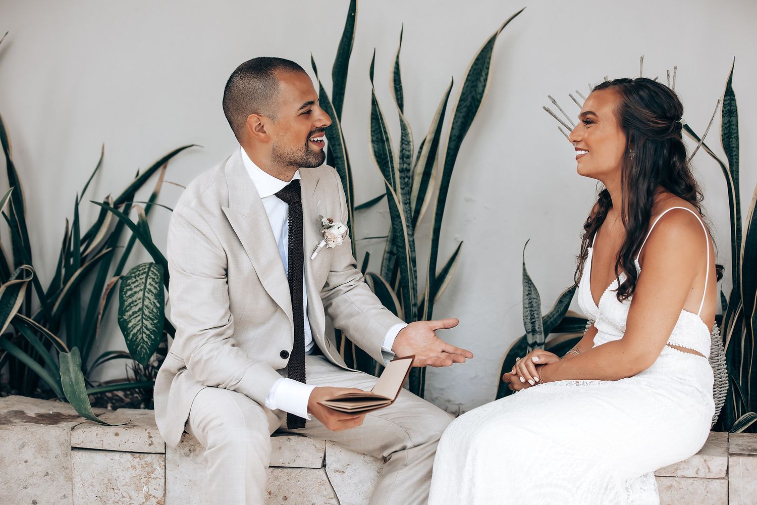 Wedding couple interacting in front of a white wall with decorative plants.