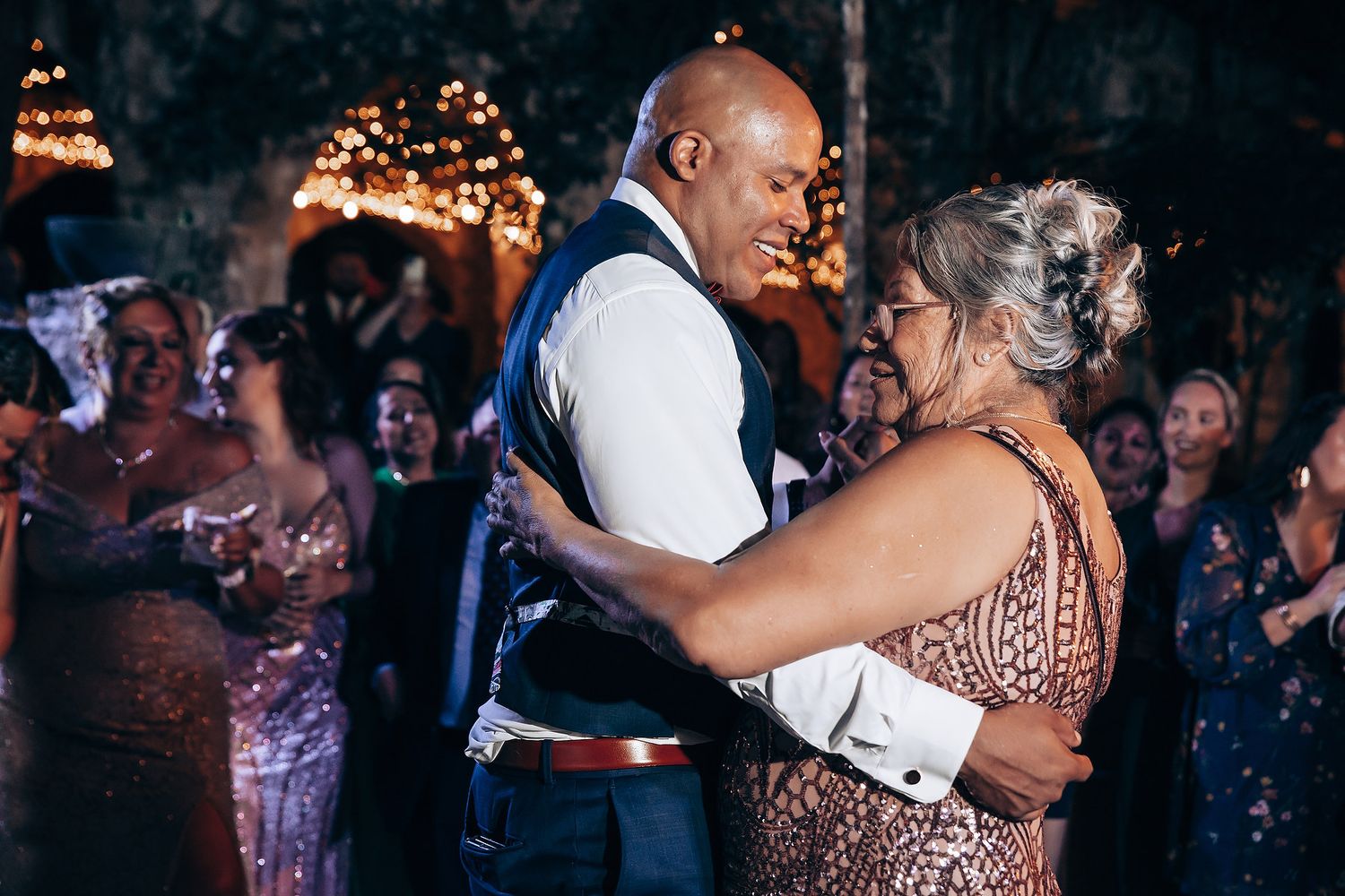 A couple shares a romantic dance moment at an elegant evening event with warm lighting and guests in the background.