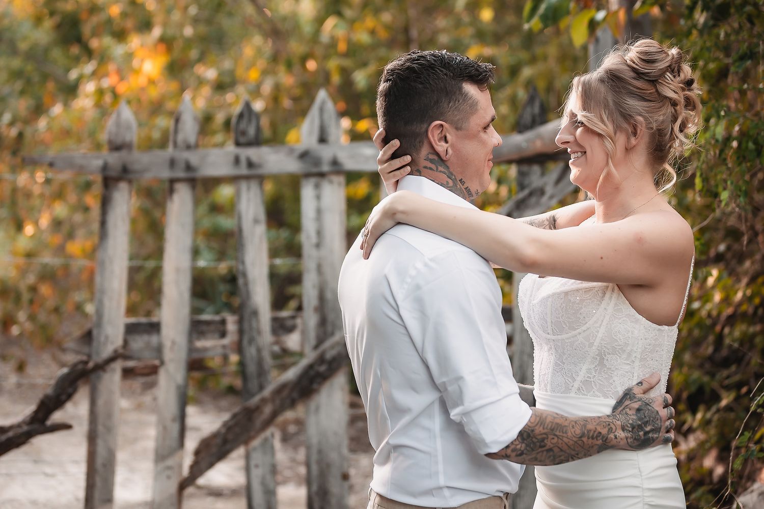 A bride and groom share a tender moment outdoors near a rustic wooden fence, surrounded by autumn foliage.