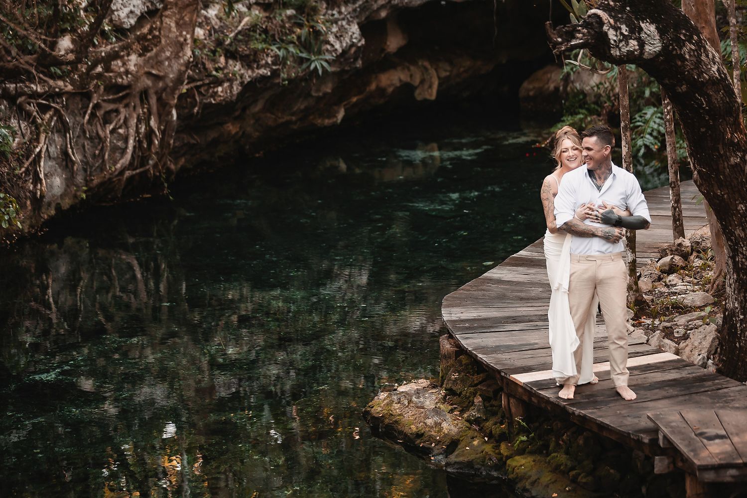 Romantic couple standing on a wooden dock over a crystal-clear cenote with lush green surroundings.