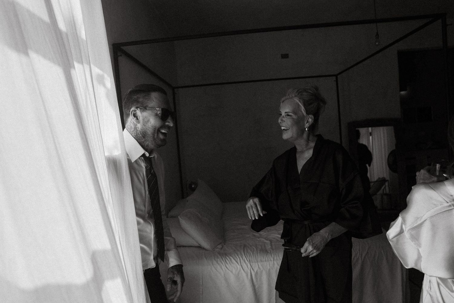Black and white photo of a man and woman in elegant attire conversing in a softly lit bedroom.