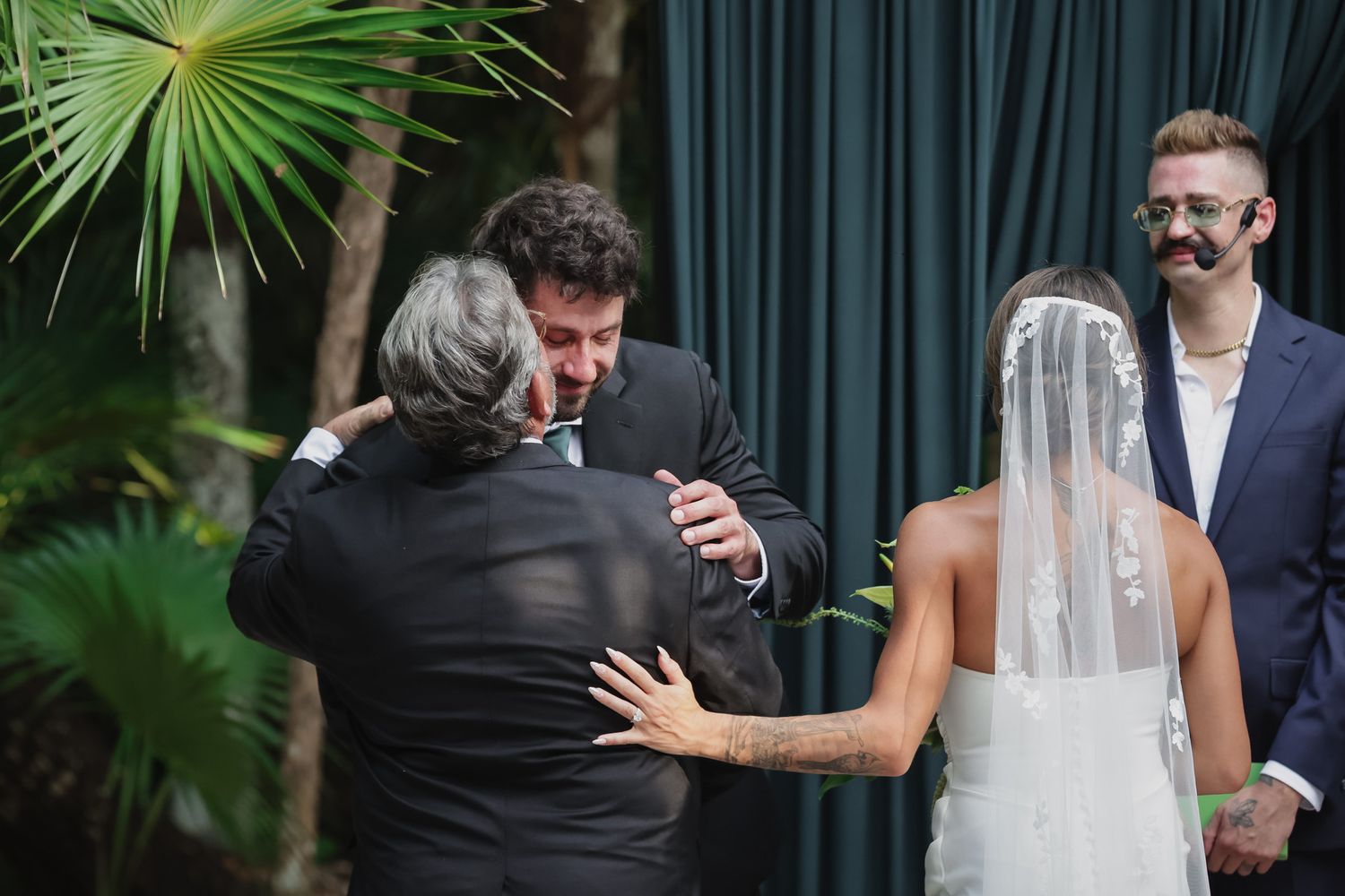 Groom embracing family member during outdoor wedding ceremony with bride in veil nearby.