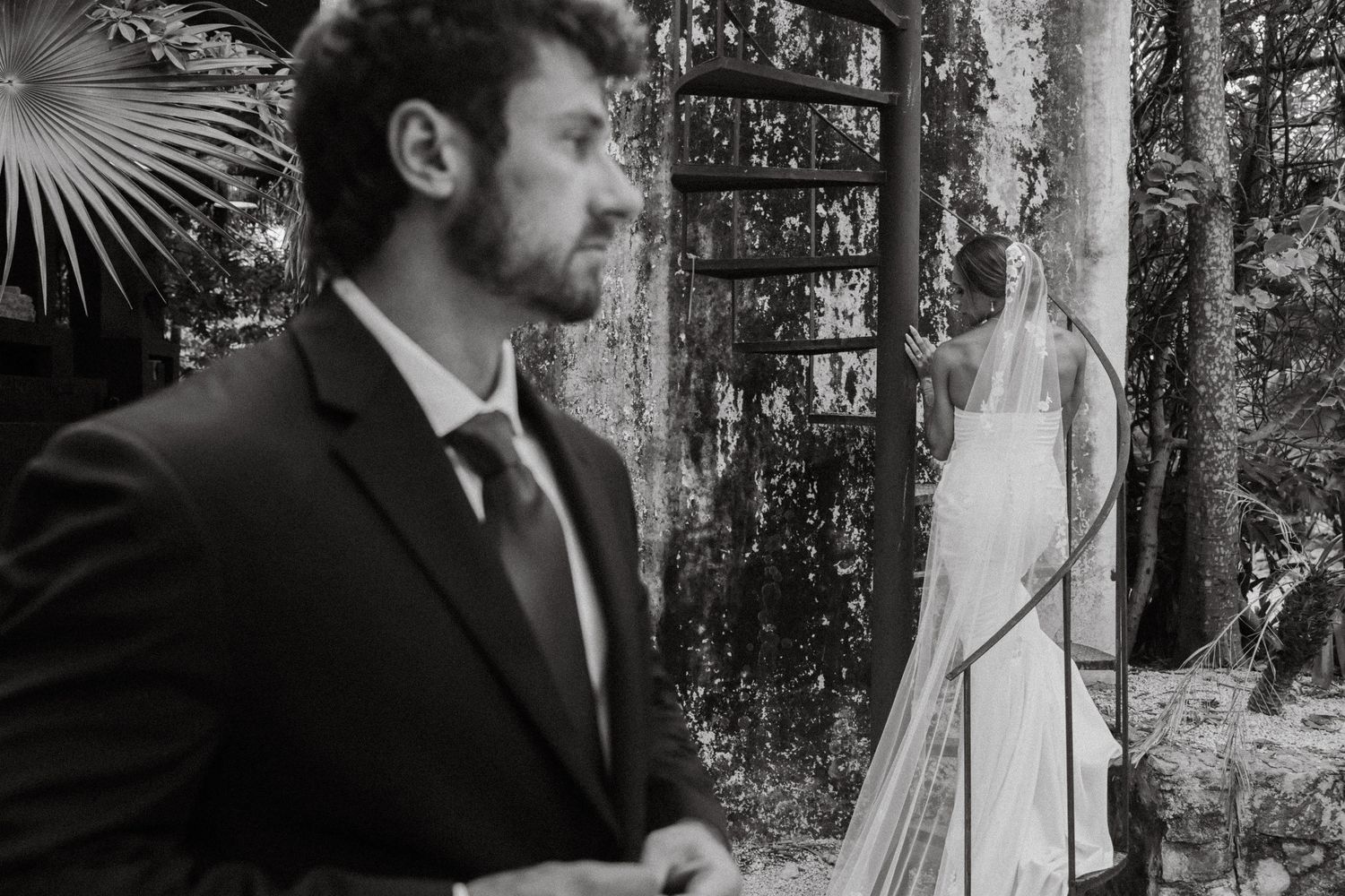 Black and white wedding photo of groom in suit with bride in white gown standing near ornate gate in lush garden.