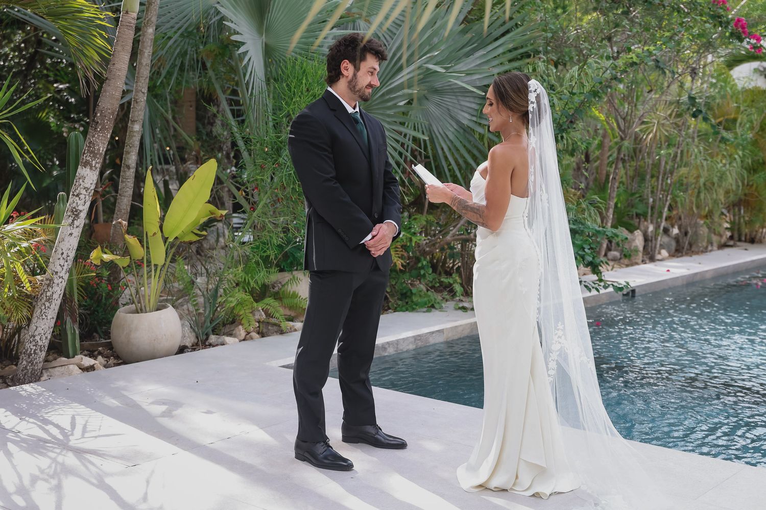 Couple exchanging wedding vows outdoors by a pool, bride in white gown with veil, groom in black suit.