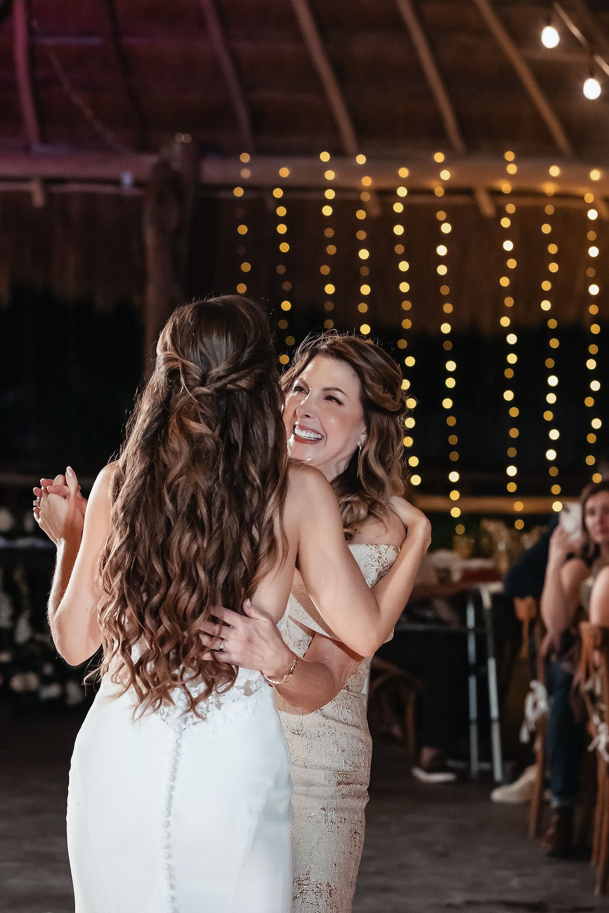 Two brides share a joyful embrace while dancing at their rustic barn wedding reception with warm string lights glowing behind them.