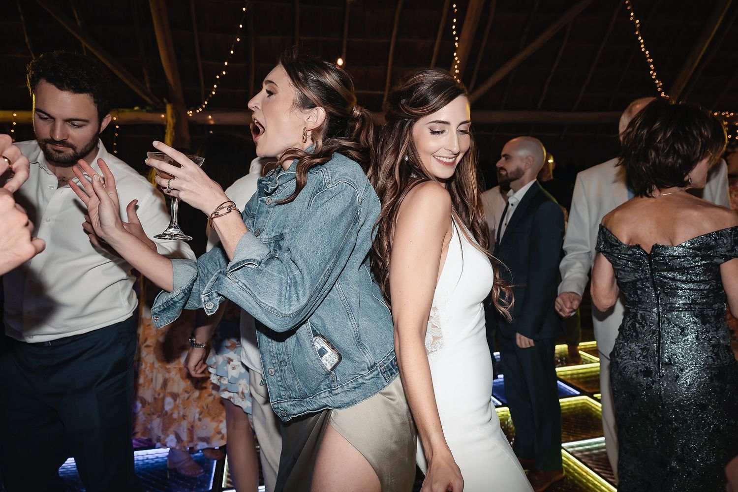 Guests dancing joyfully at a wedding reception, bride in white gown smiling with friend in denim jacket.