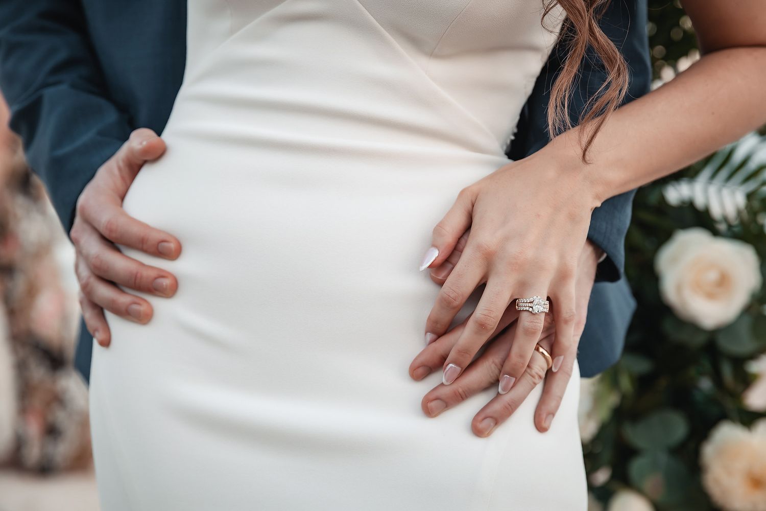 Bride and groom hands on white wedding dress, showing diamond engagement ring and wedding band near floral arrangement.