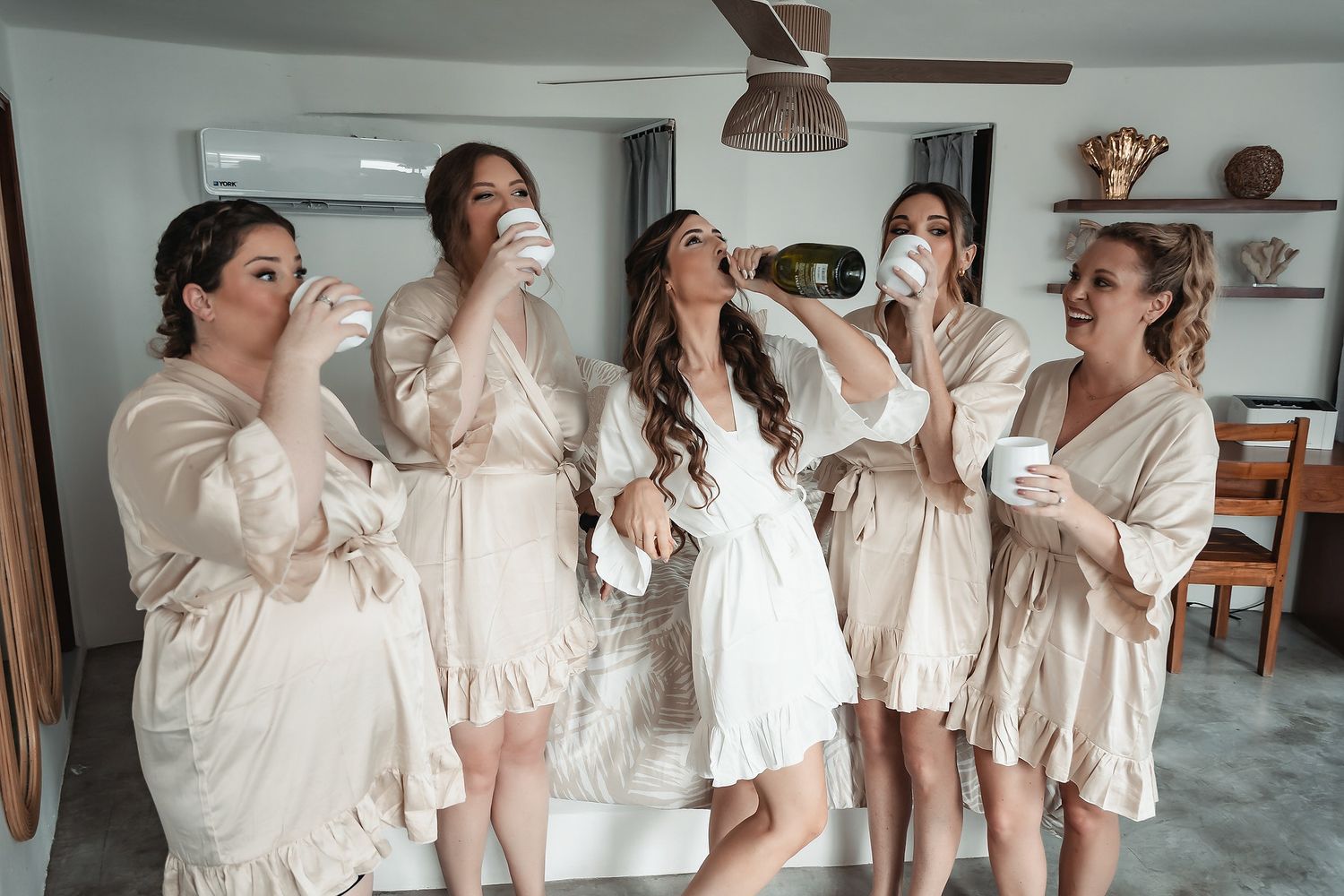 Bridesmaids in matching satin robes toasting with coffee cups alongside bride in white robe during wedding morning preparations.