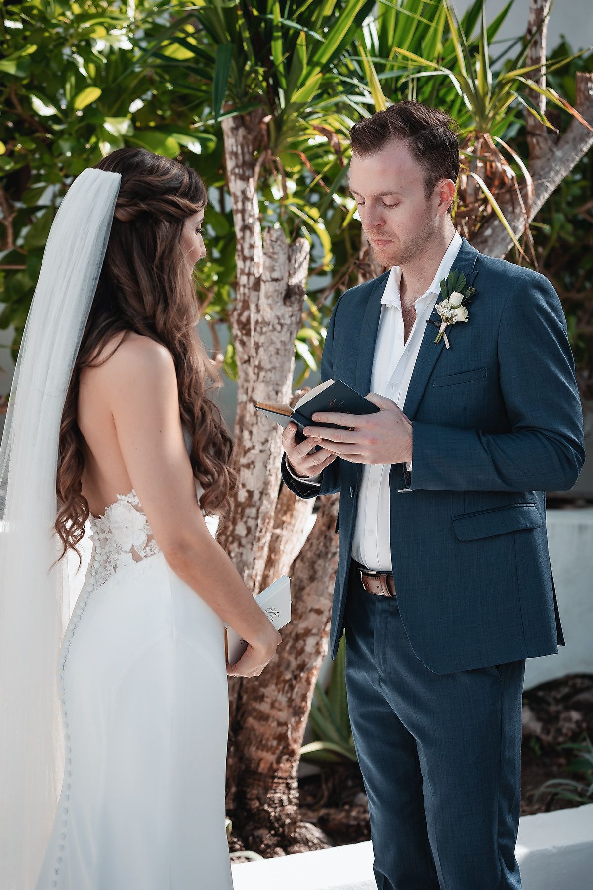 Groom in navy suit reading vows from book to bride in lace wedding dress outdoors.