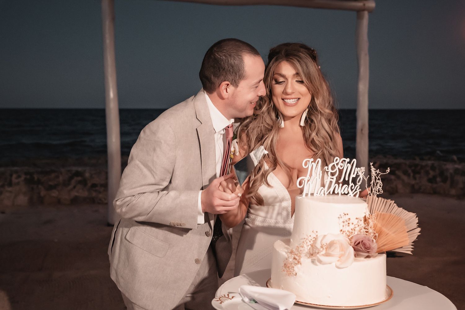 Bride and groom cutting wedding cake on beach at night, wearing elegant attire with Mr. and Mrs. cake topper.