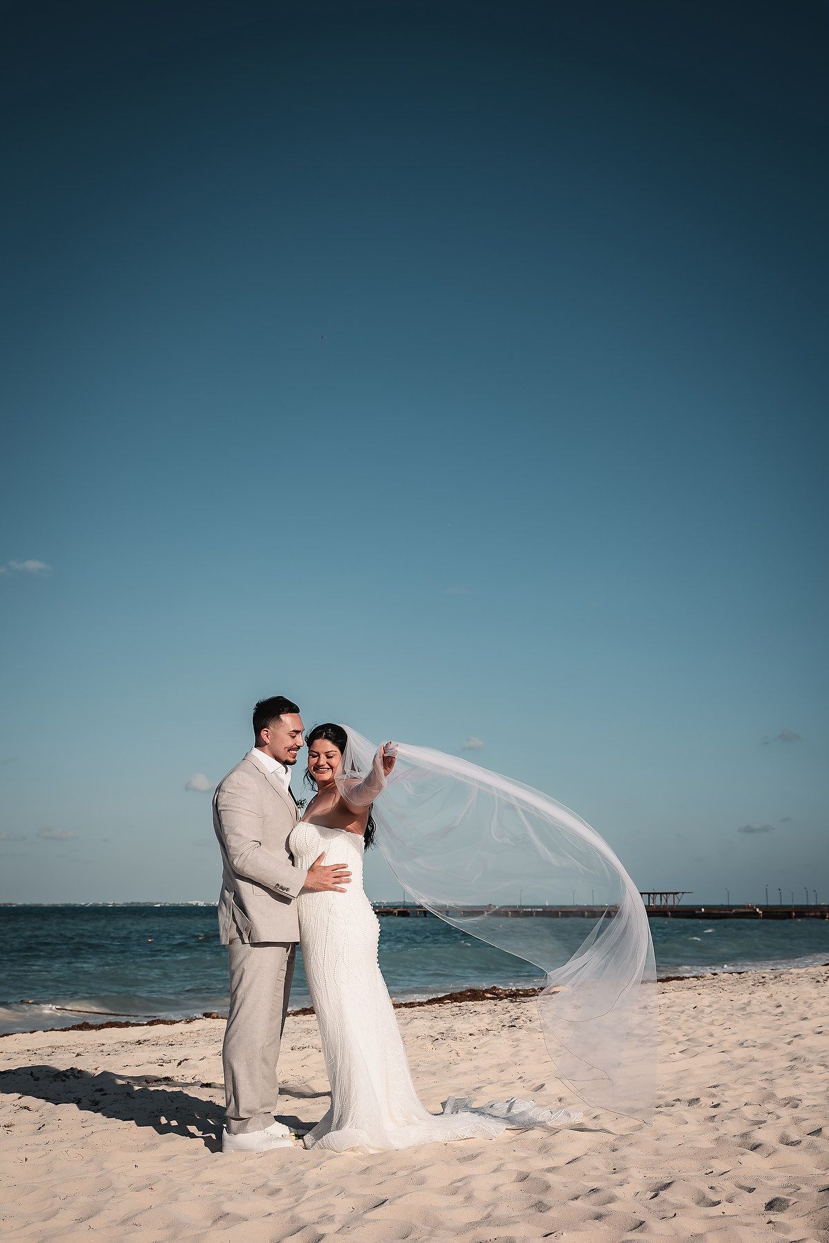 Bride and groom embrace on a sunny beach, her veil flowing in the wind, with turquoise ocean waters in the background.