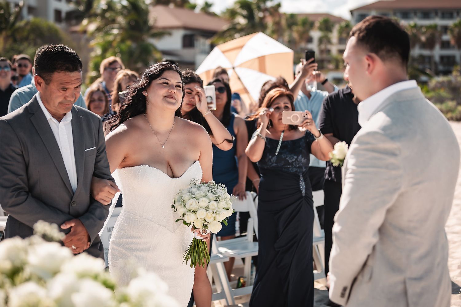 Bride walks down the aisle holding white bouquet at outdoor beach wedding ceremony with guests watching.