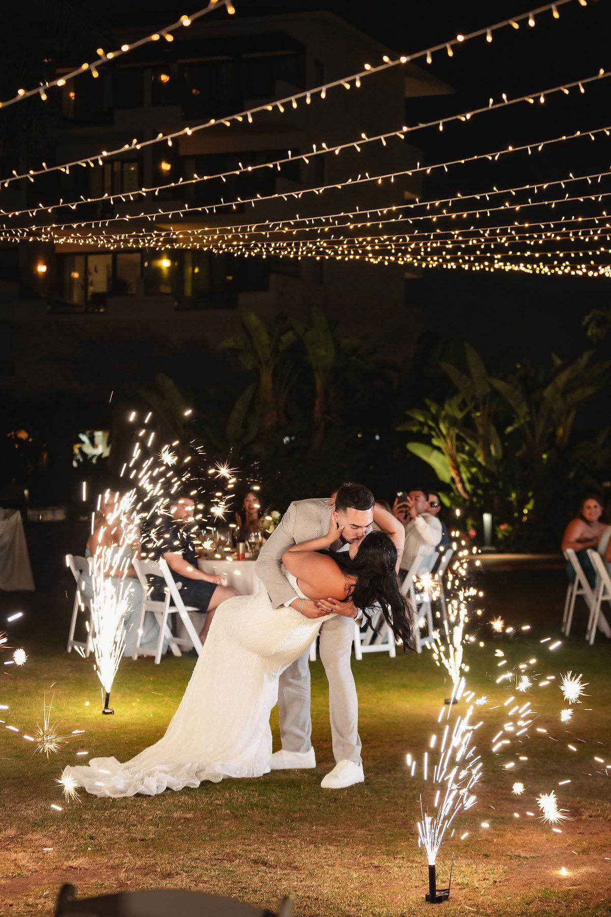 Bride and groom share a dip kiss surrounded by sparkling fountains under string lights at outdoor wedding reception.