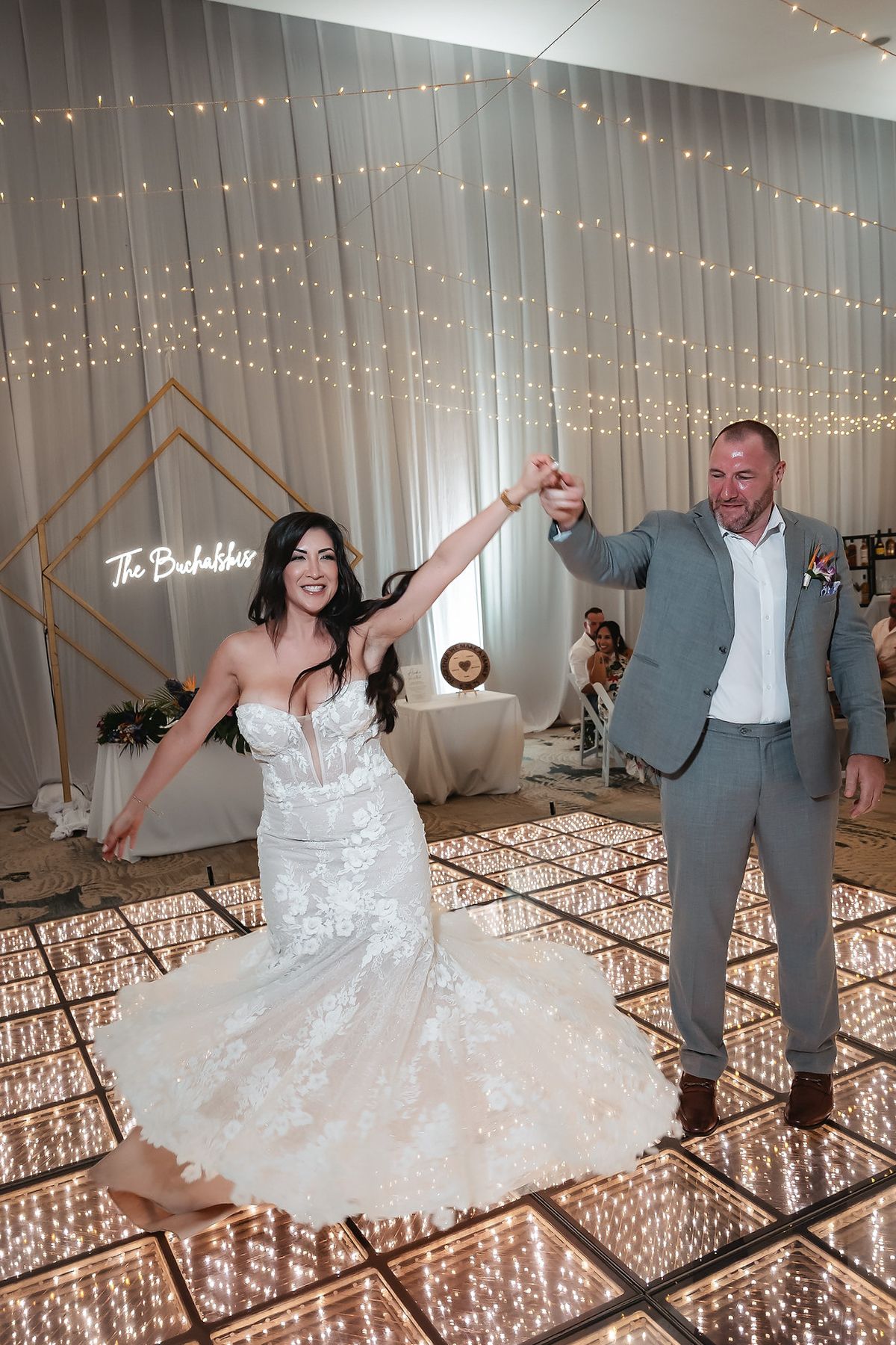 Bride and groom share first dance on illuminated dance floor at elegant wedding reception with fairy lights backdrop.