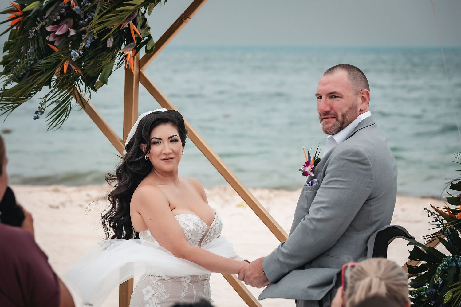 Bride and groom holding hands at a beach wedding ceremony with ocean backdrop and tropical floral arch.