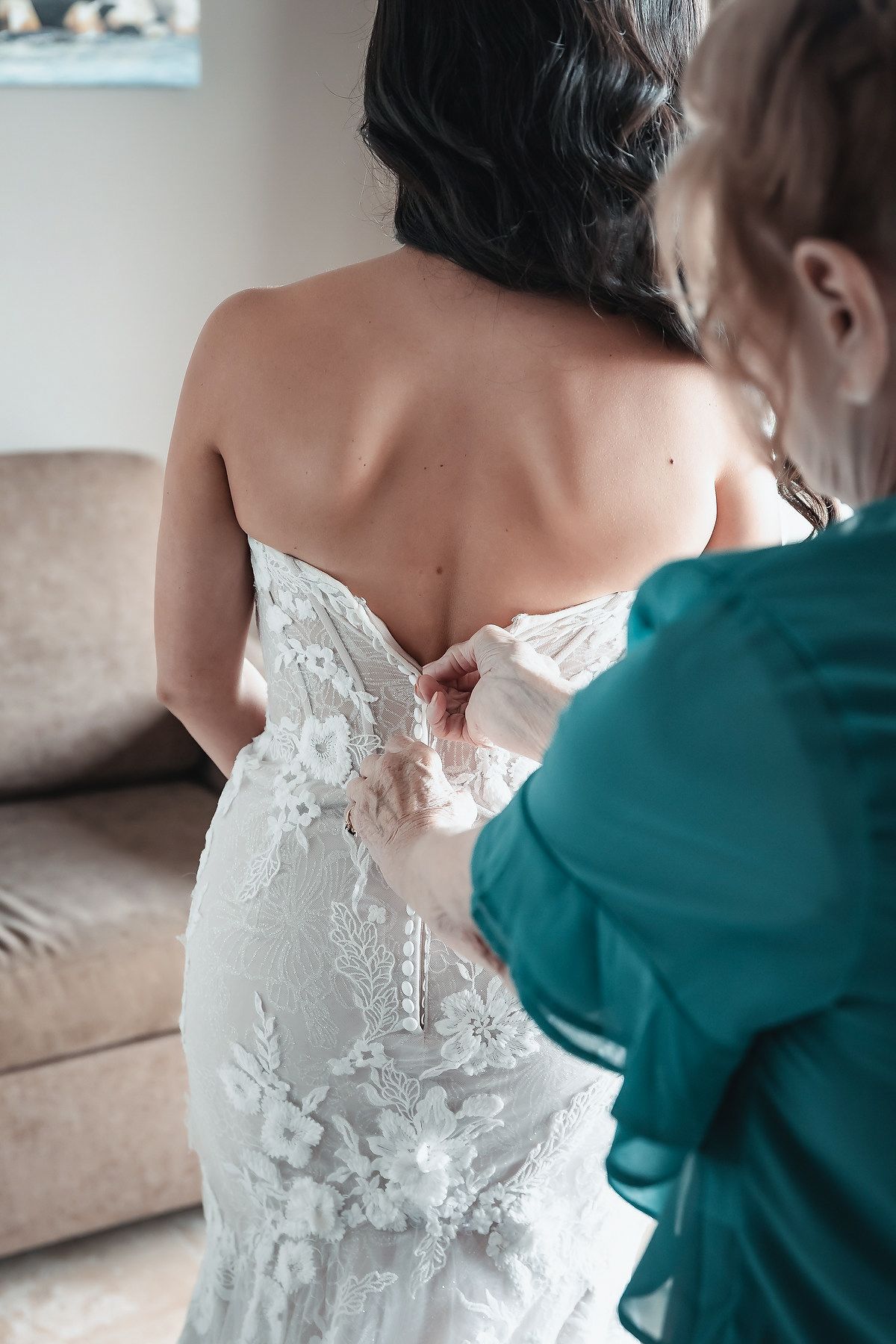 Bridesmaid buttoning back of lace wedding dress on bride before ceremony.