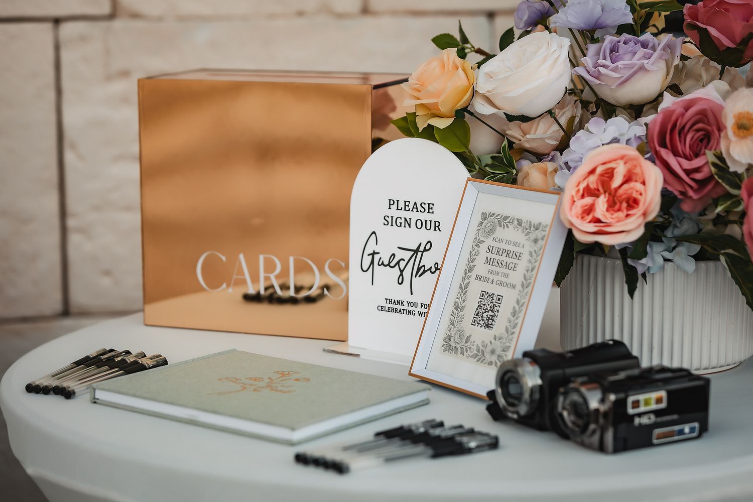 Wedding guest book table with cards box, floral arrangement, sign, camera, and pens on white table.