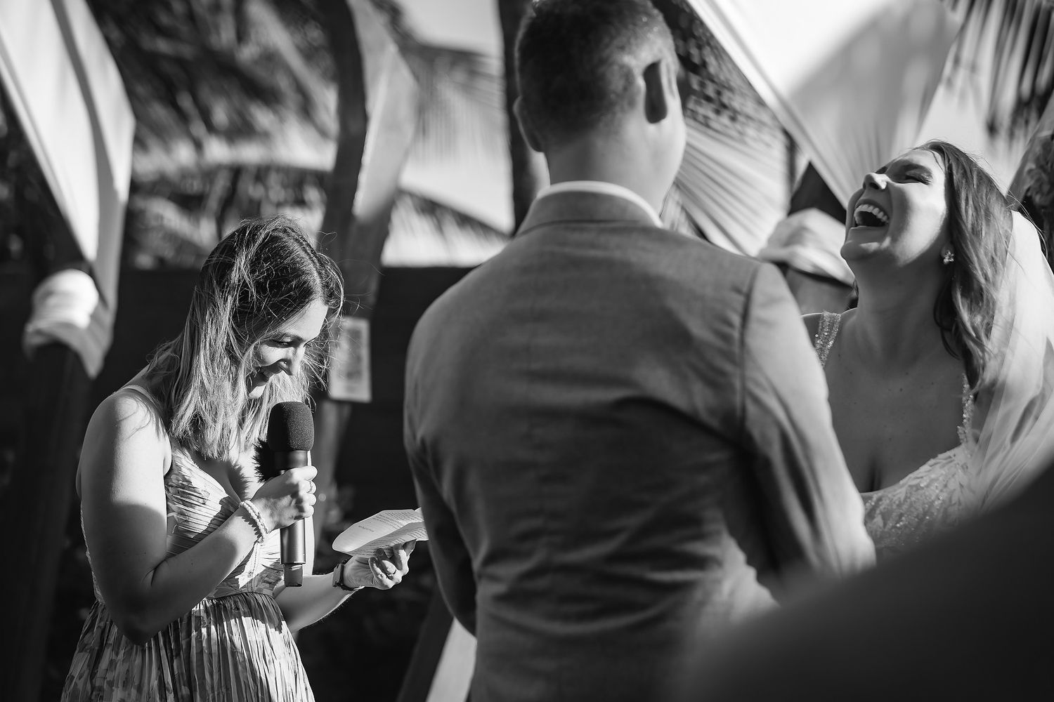 Black and white wedding photo of bride laughing joyfully with groom and bridesmaid during outdoor ceremony.