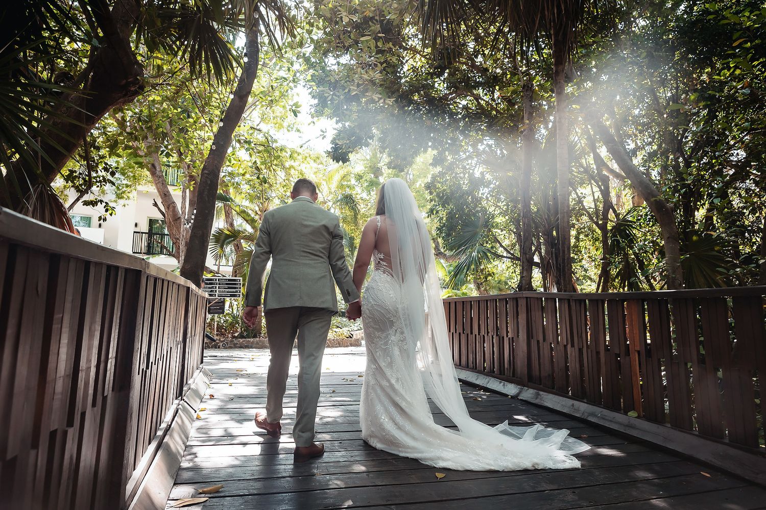 Bride and groom walking hand-in-hand on a sunlit wooden bridge surrounded by lush green trees.