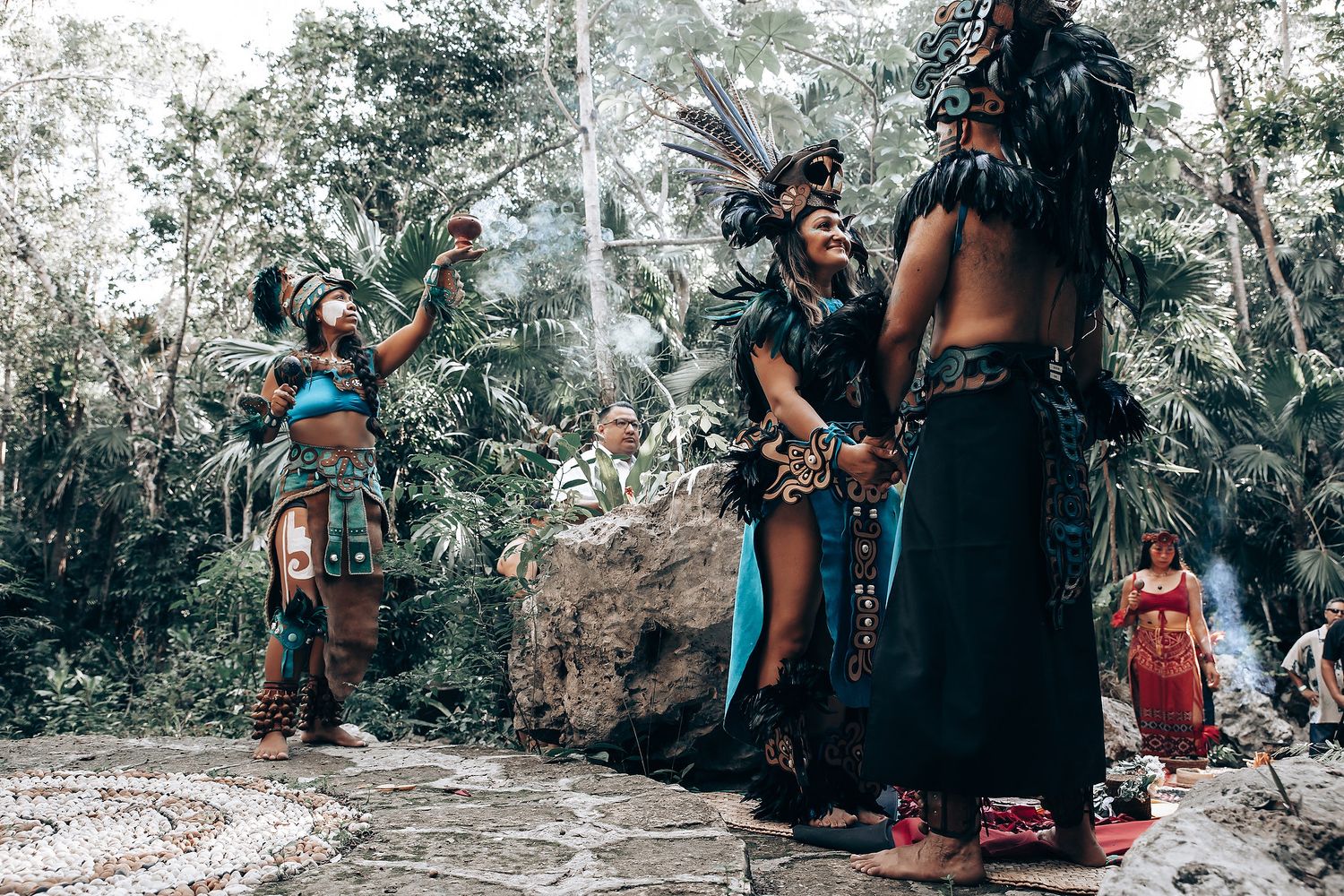 Group of performers in traditional Mayan ceremonial costumes conduct an outdoor ritual on stone ruins in a jungle.