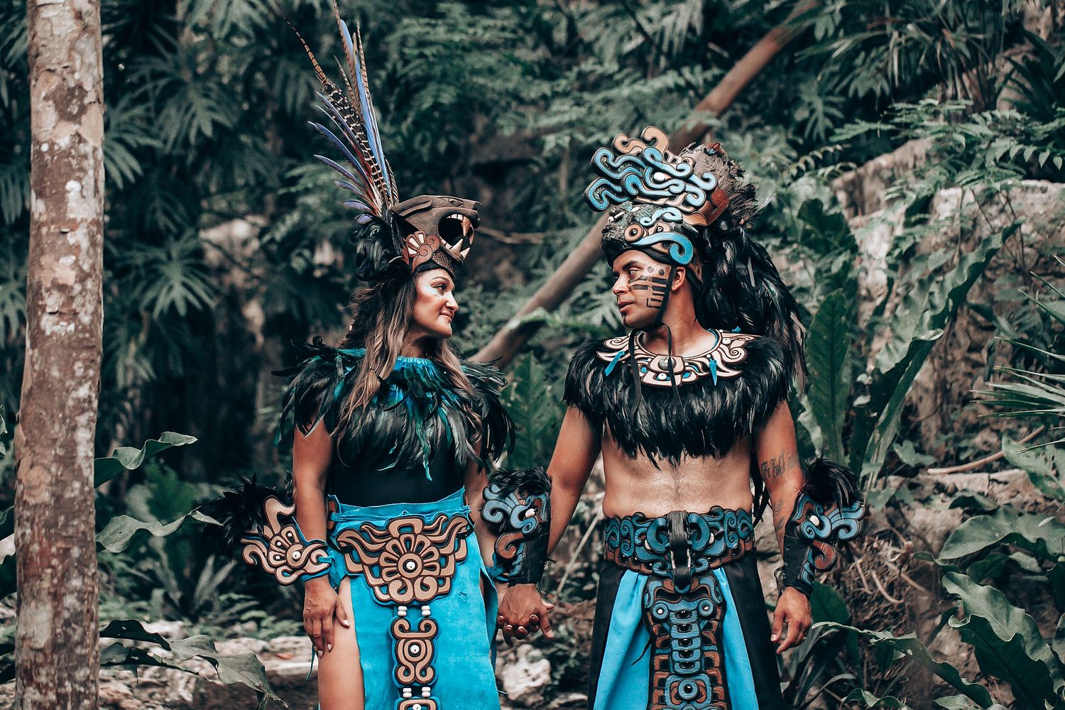 Two performers in matching turquoise Mayan ceremonial outfits with feathered headdresses facing each other in jungle.
