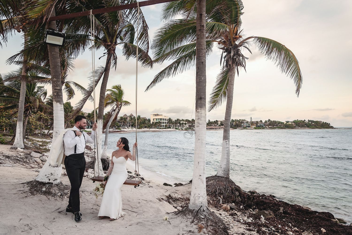 Bride sits on a beach swing as groom stands beside her under palm trees at sunset.